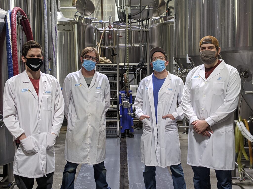 Four people in lab coats and masks in front of metal tanks