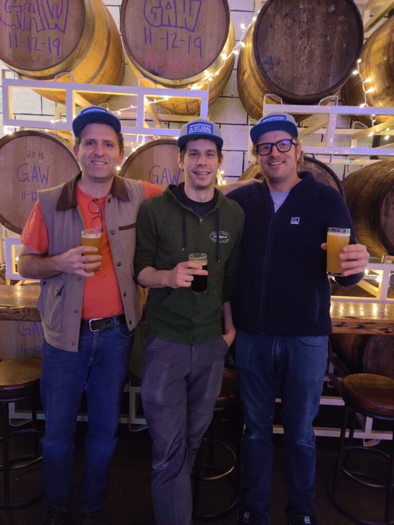Three men hold beers in front of storage barrels at a brewery